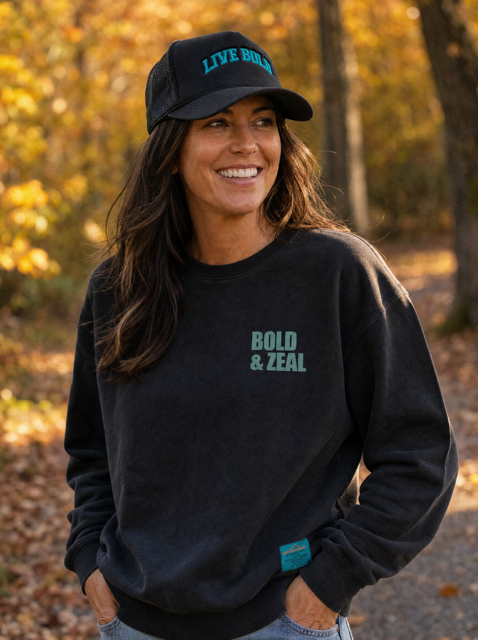 A woman outdoors in a wooded area with autumn foliage, wearing a black sweatshirt with 'BOLD & ZEAL' printed in teal on the chest, a matching black baseball cap with 'LIVE BOLD' in teal, and light blue jeans.